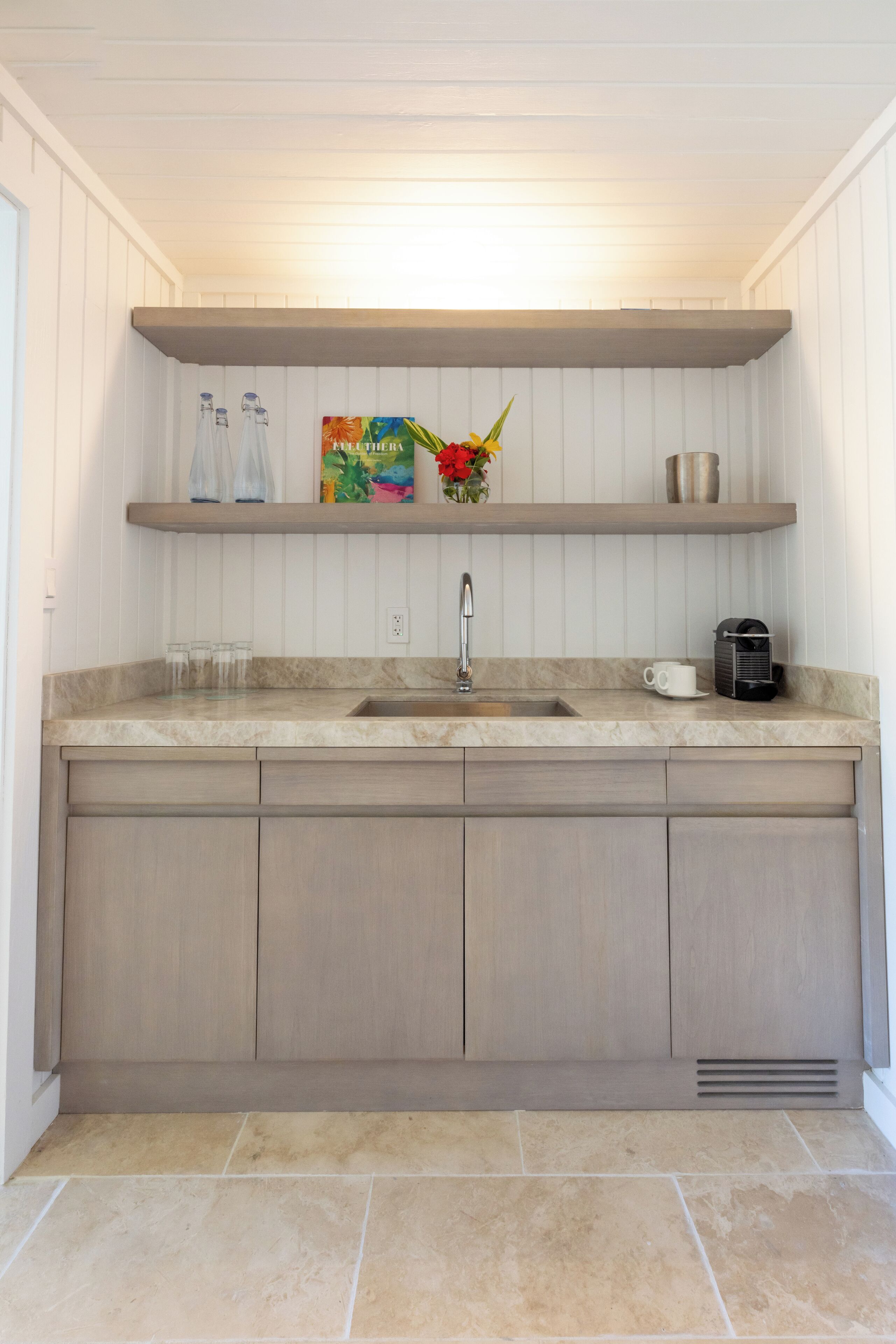 A kitchen with a double sink, wooden cabinets, and a countertop. There are two glass bottles, a coffee maker, and a decorative item on the shelf.