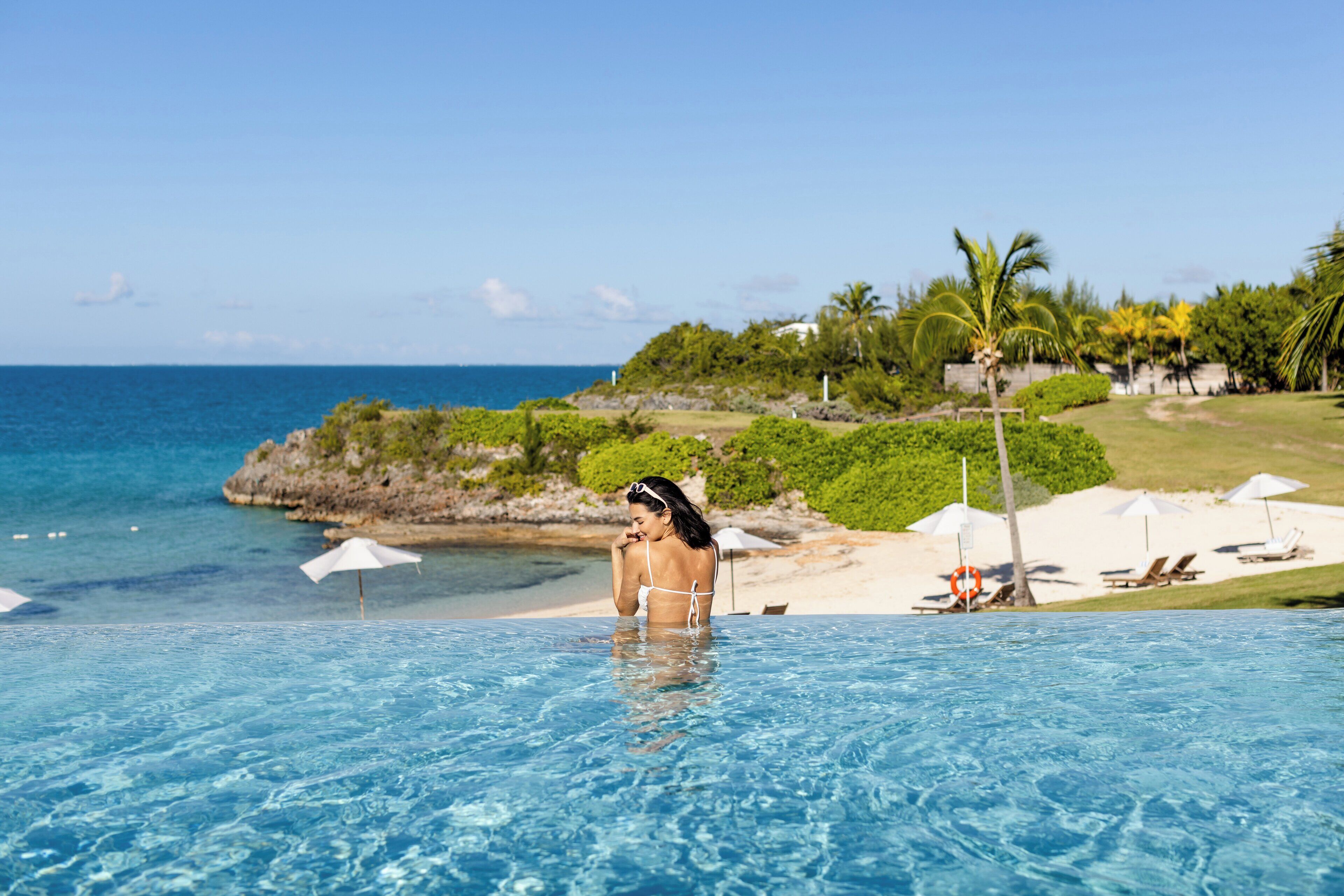 A pool with clear blue water, a woman in a bikini, and a beach with umbrellas and lounge chairs in the background.