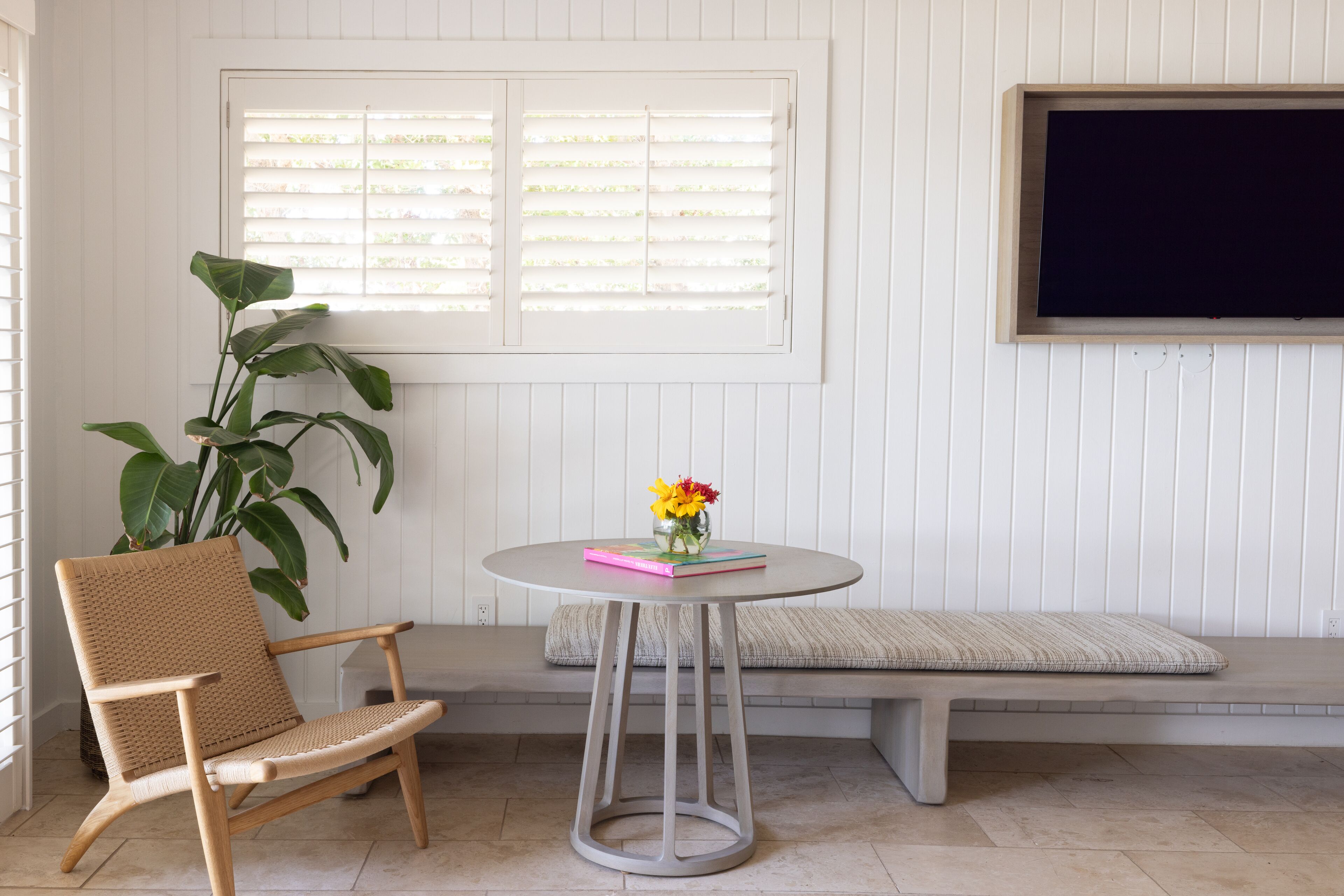 A room with a white paneled wall, a bench, a wicker chair, a round table with a vase of flowers, and a television mounted on the wall.