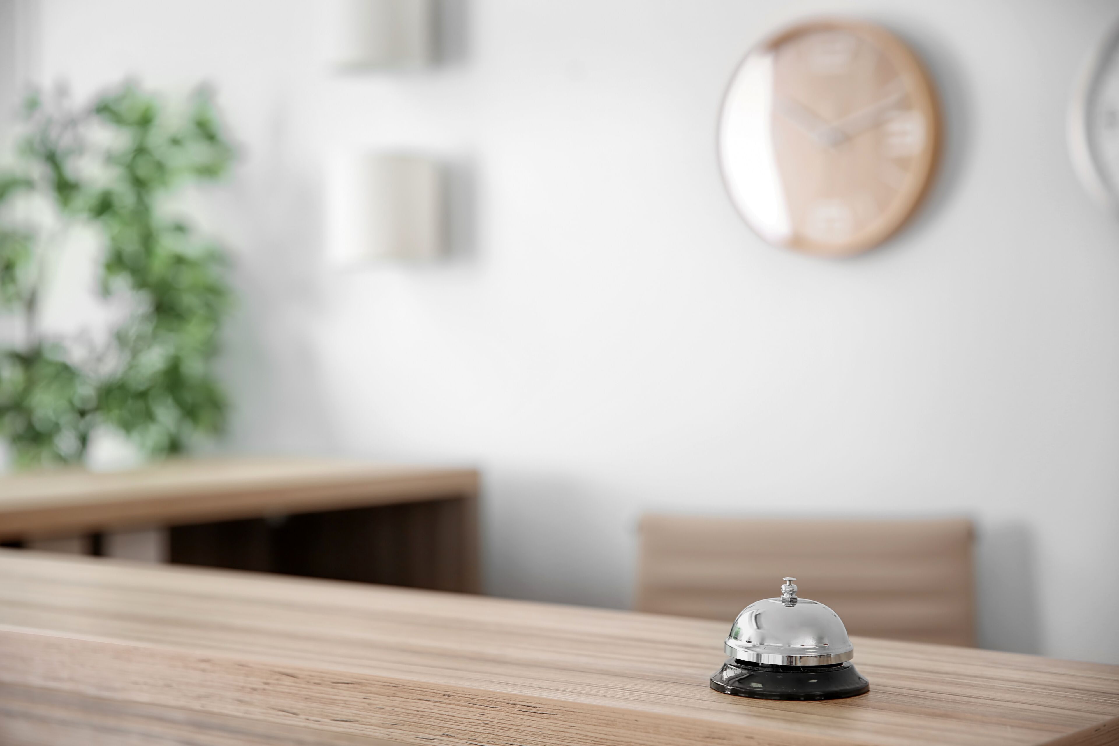 Une table en bois sur laquelle se trouve une cloche noire et argentée, une horloge en bois accrochée au mur et une plante floue en arrière-plan.
