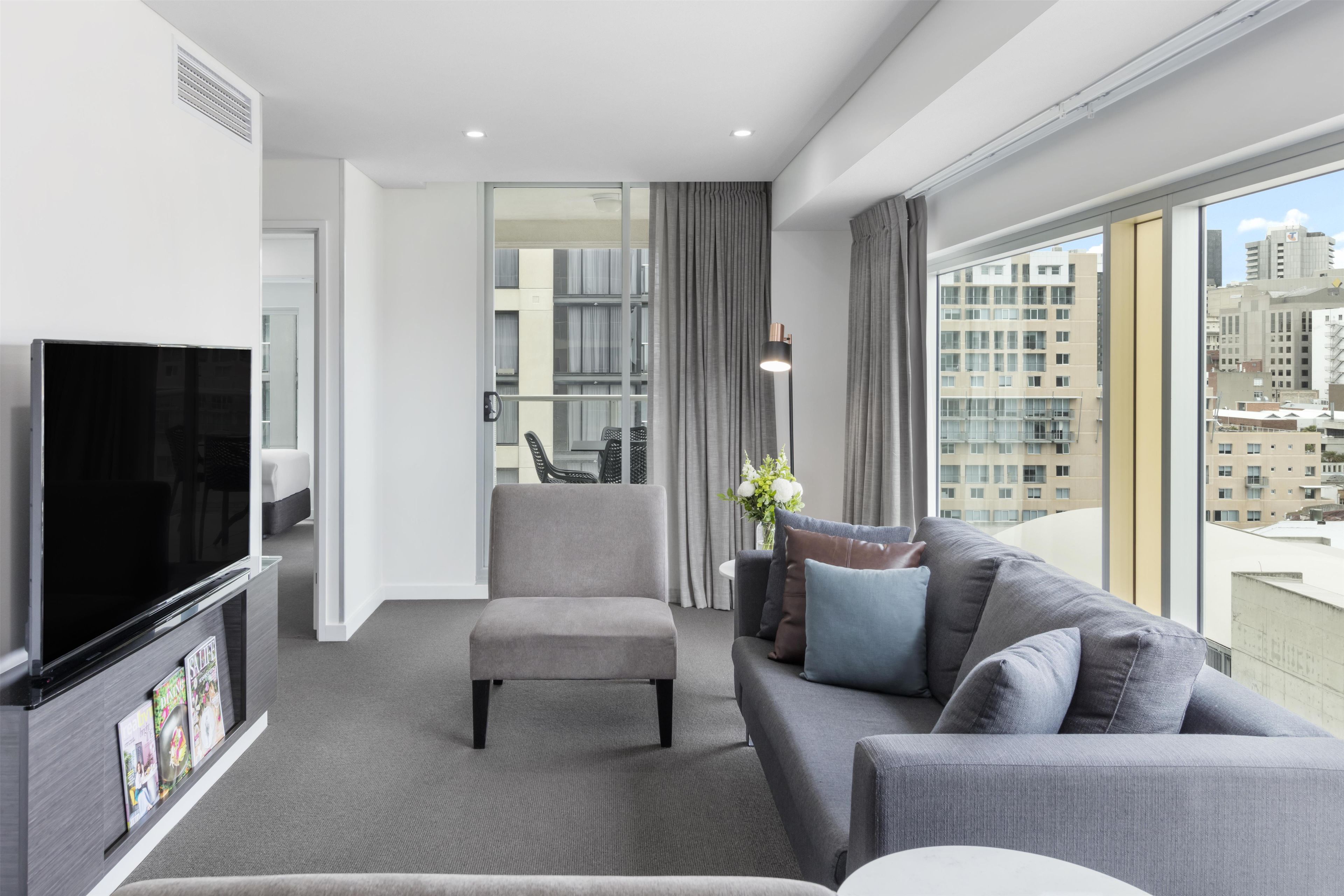 Et moderne hotelværelse med et fladskærms-tv, en grå sofa, en mindre lænestol og et stort vindue med udsigt over byen.