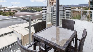 Un balcón con mobiliario de mimbre y una mesa de vidrio con vistas a un paisaje urbano que incluye edificios y un estadio.