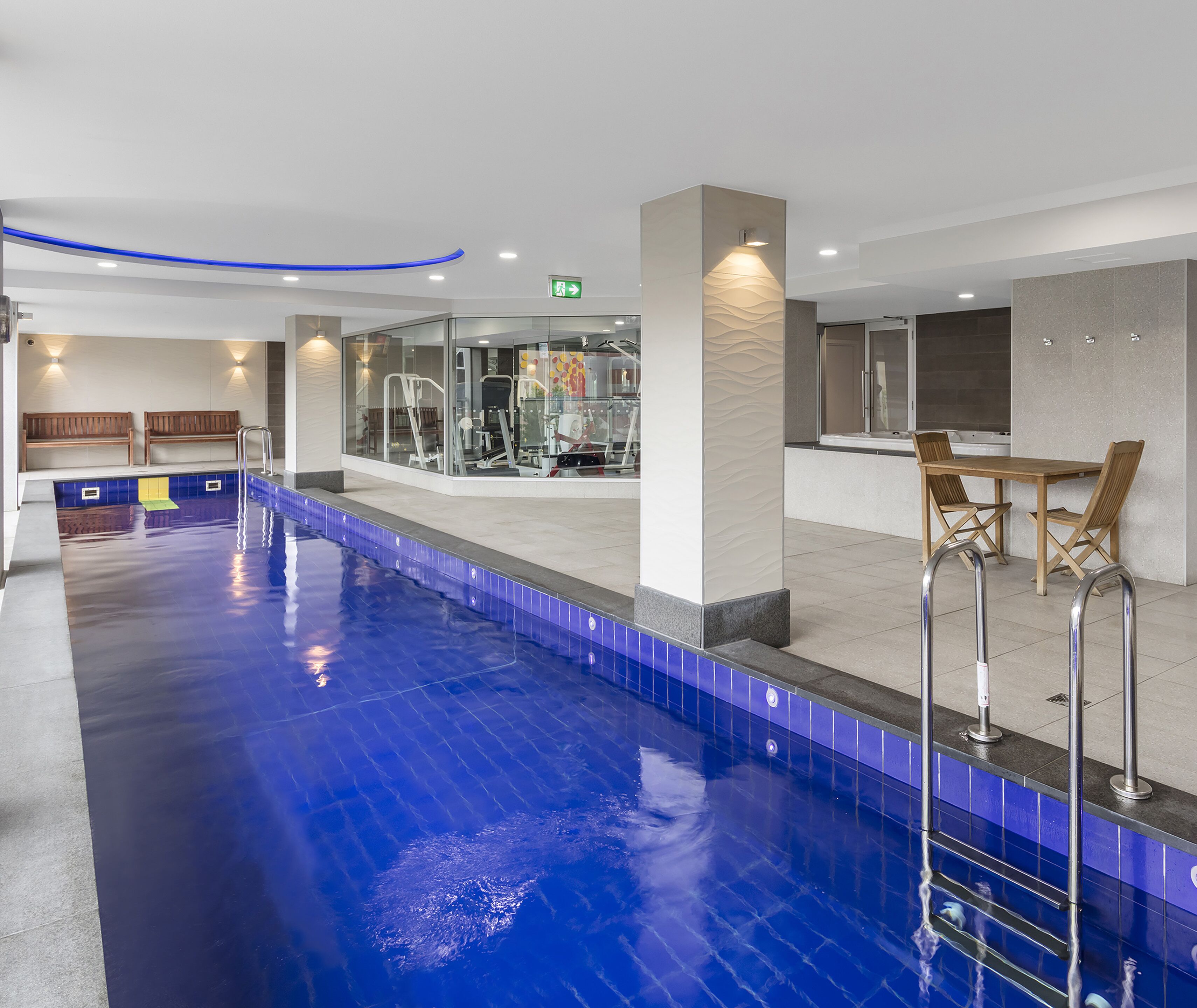 A swimming pool with a blue tiled edge, a fitness area with exercise machines, and a seating area with wooden chairs and a table.