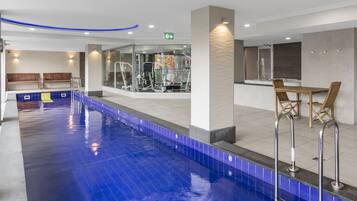 A swimming pool with a blue tiled edge, a fitness area with exercise machines, and a seating area with wooden chairs and a table.