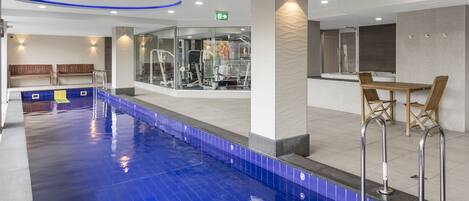 A swimming pool with a blue tiled edge, a fitness area with exercise machines, and a seating area with wooden chairs and a table.