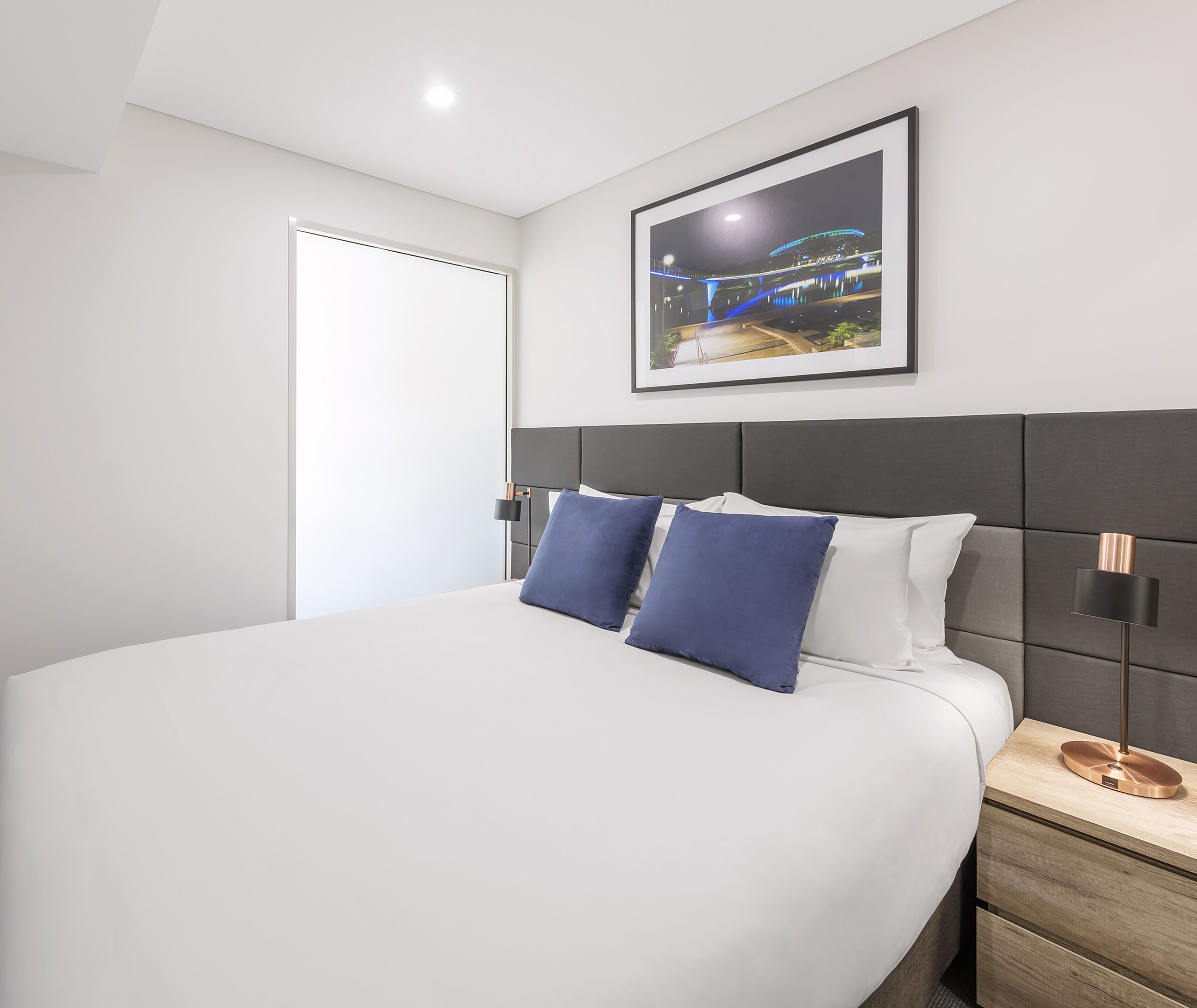 A modern hotel room with a large bed, a framed picture on the wall, and a nightstand with a lamp.