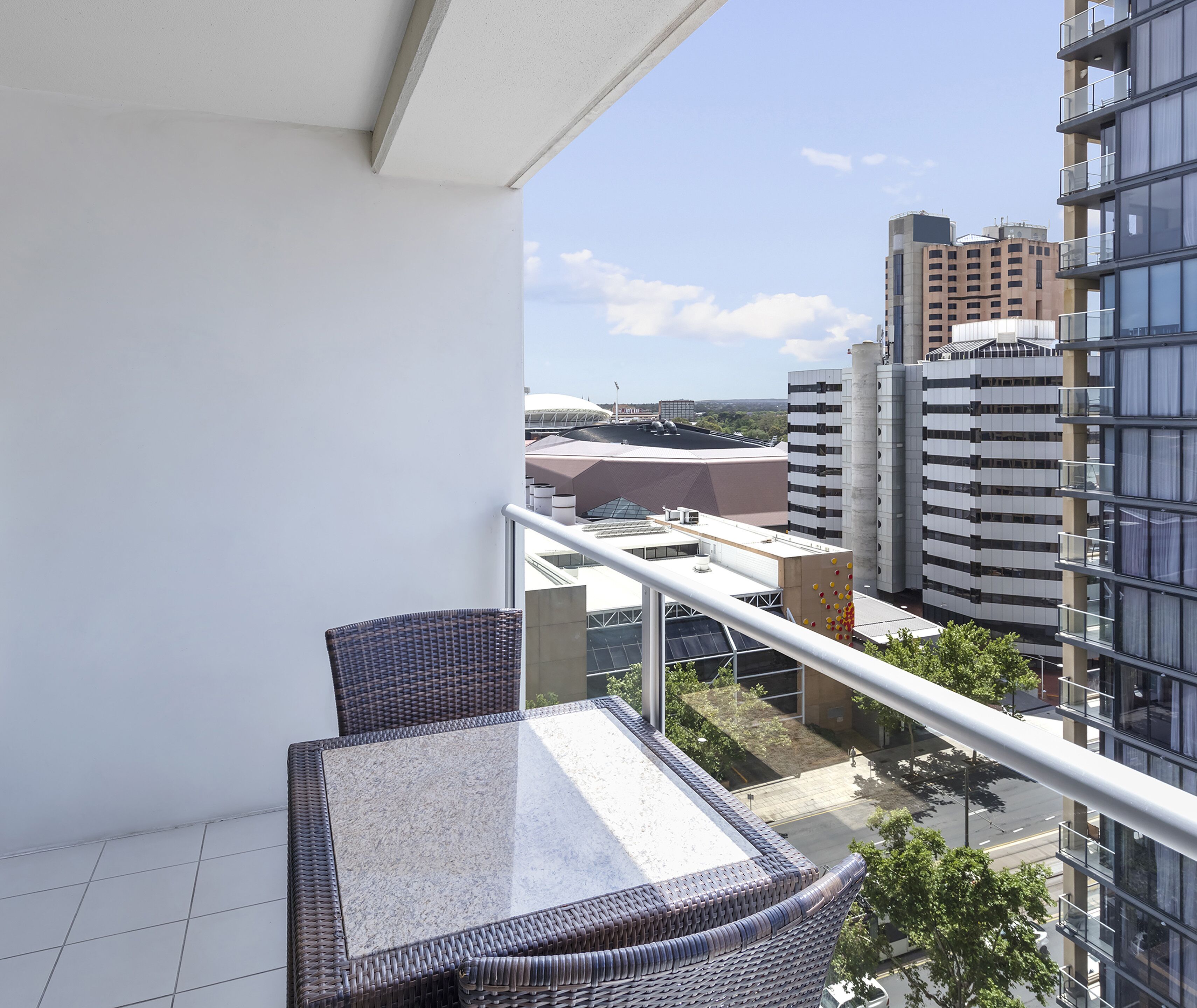 A balcony with wicker furniture and a glass table, overlooking a cityscape with modern buildings and a clear sky.