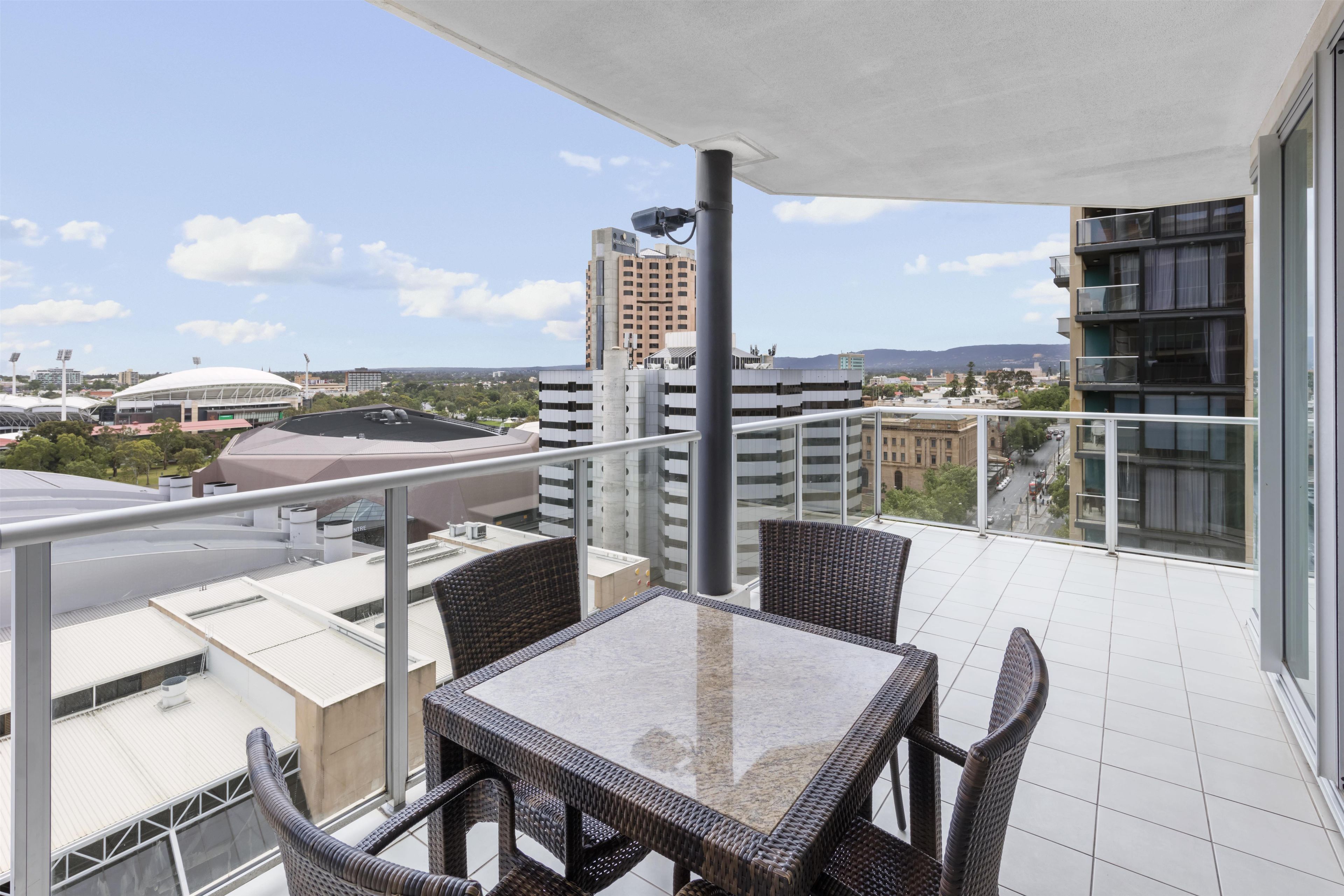 Un balcón con mesa y sillas, con vista a un paisaje urbano que incluye edificios y un estadio.