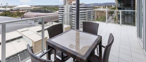 Un balcón con mesa y sillas, con vista a un paisaje urbano que incluye edificios y un estadio.