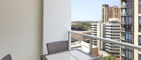 A balcony with wicker furniture and a glass table, overlooking a cityscape with high-rise buildings.