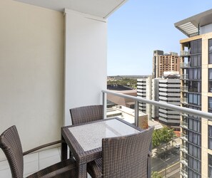A balcony with wicker furniture and a glass table, overlooking a cityscape with high-rise buildings.