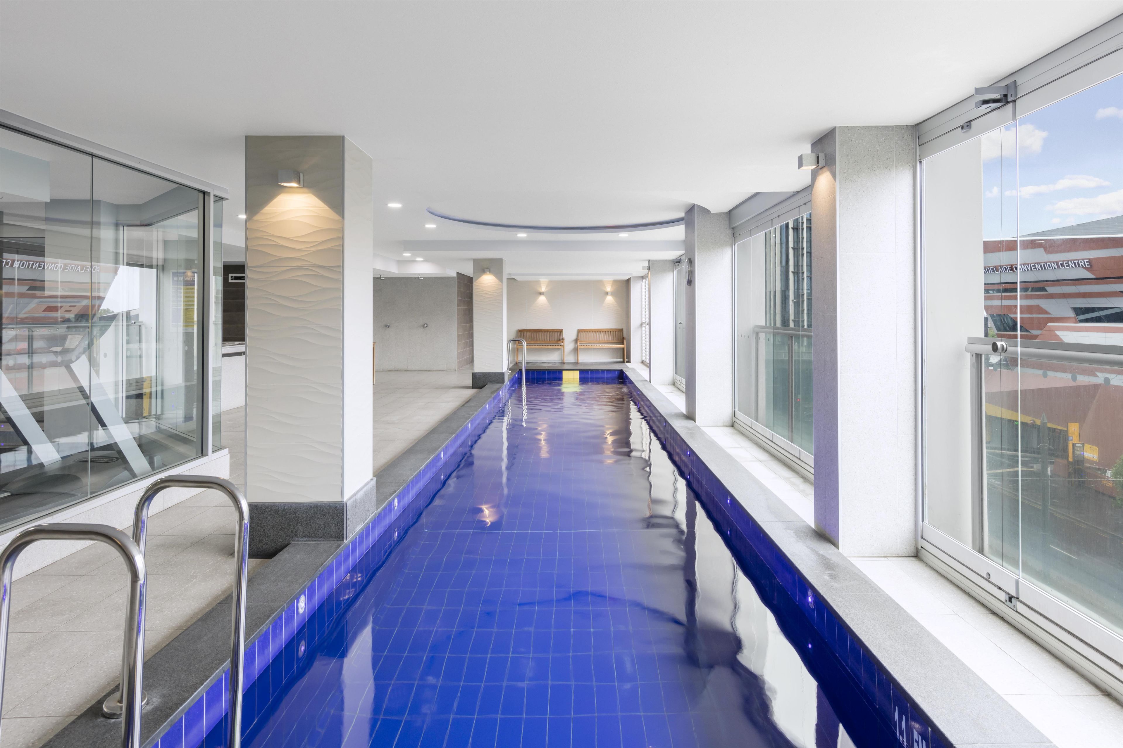 A modern indoor swimming pool with clear glass walls, a blue tiled floor, and a bench along the side.