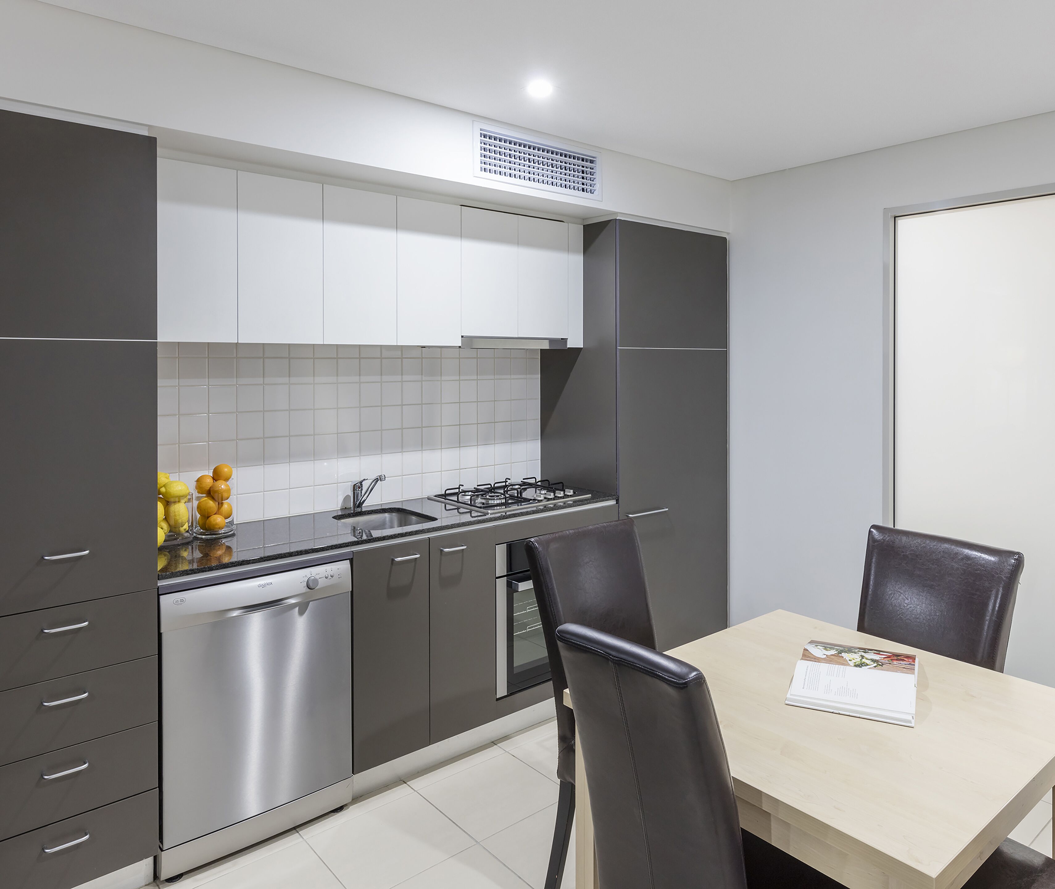 A modern kitchen with grey cabinets, a stainless steel dishwasher, and a gas stove. There is a dining table with two chairs and a magazine on it.