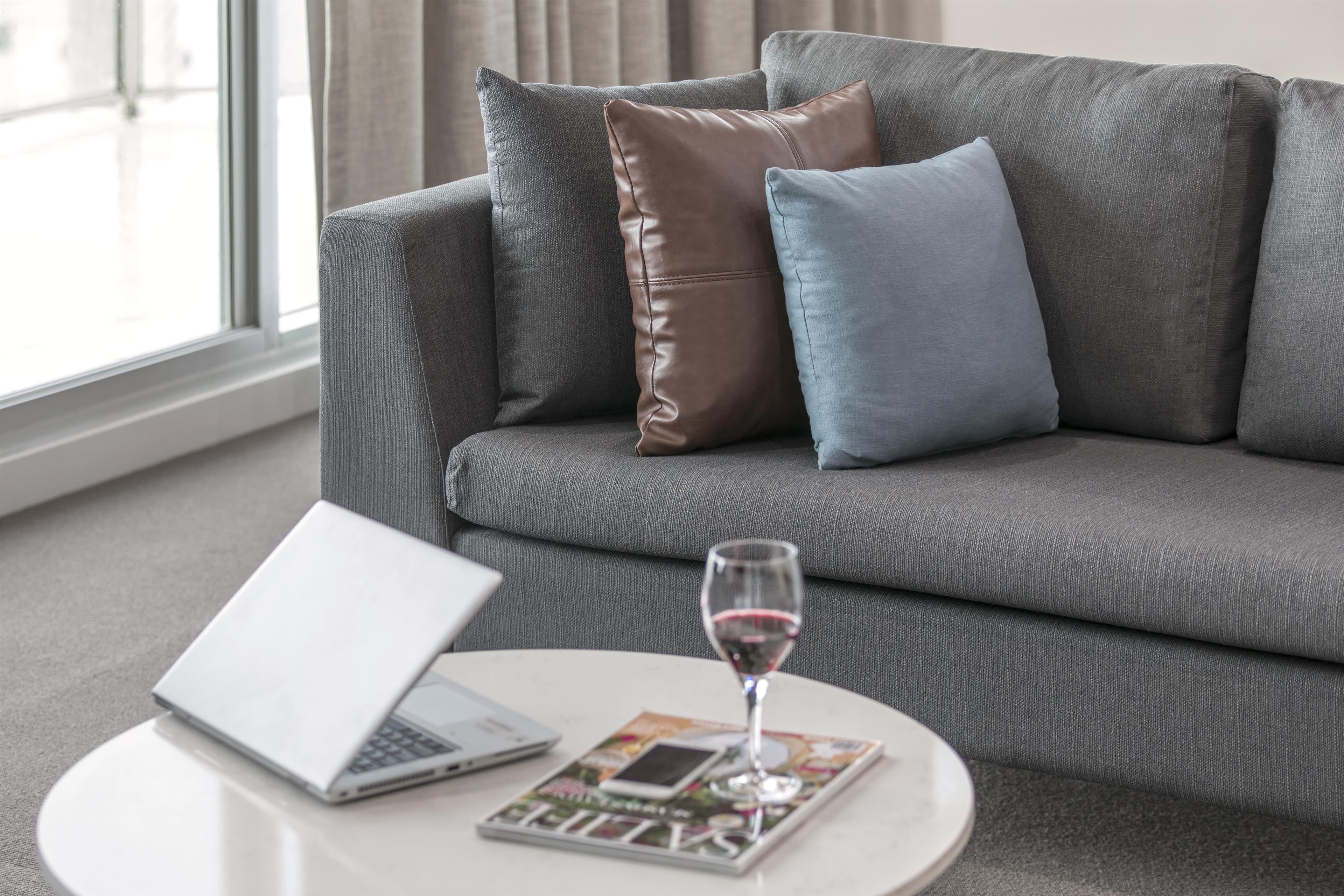 Un canapé gris avec des coussins en cuir et en tissu, un ordinateur portable, un verre de vin et un magazine sur une petite table ronde.