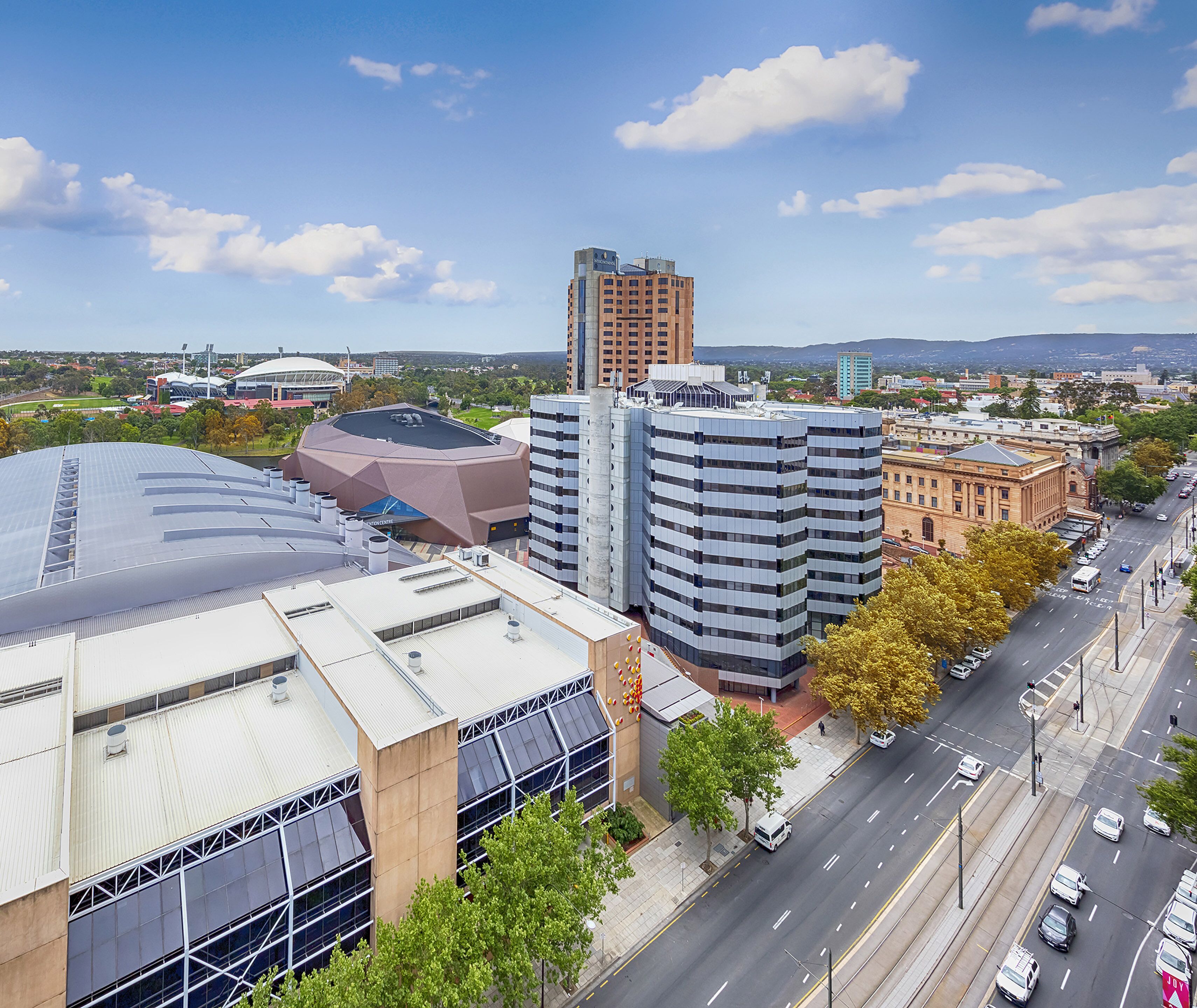 A modern building with a curved roof and large windows, surrounded by a cityscape with various other buildings and a clear sky.