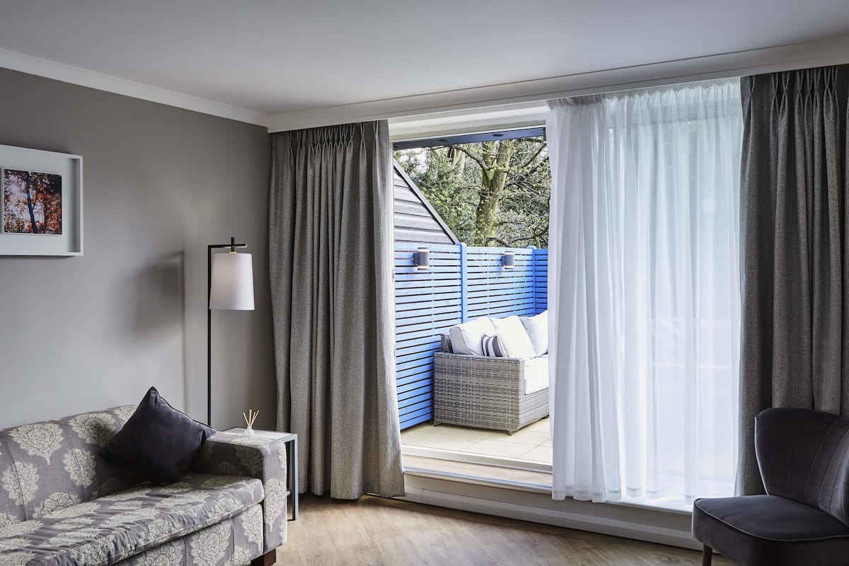 Ein modernes Wohnzimmer mit einer Couch, einem Sessel, einer Stehlampe und Blick durch eine Glastür zu einem Außenbereich mit blauem Zaun und einer Rattan Couch.