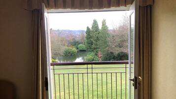 Une vue depuis un balcon doté d’une rambarde en métal, donnant sur une zone herbeuse et un plan d’eau, avec des arbres et des arbustes en arrière-plan.