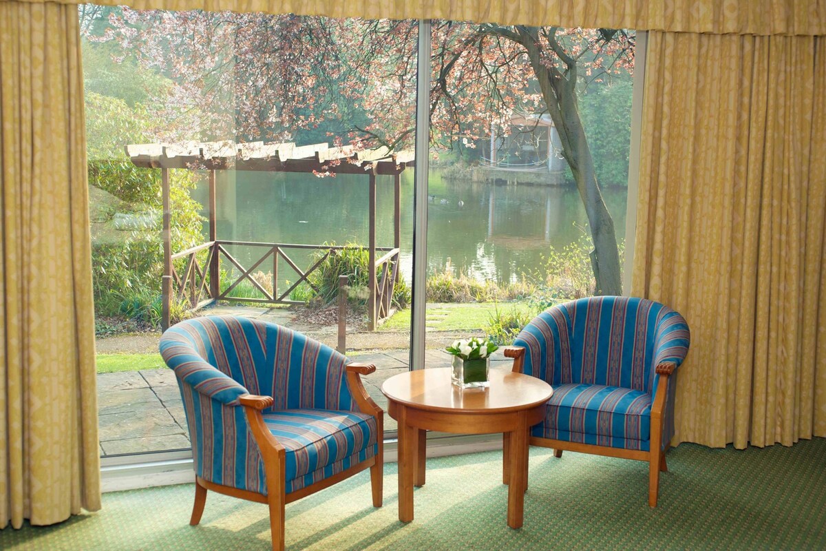 A room with two striped armchairs and a small round table, a wooden pergola, and a view of a garden and water body through large windows.