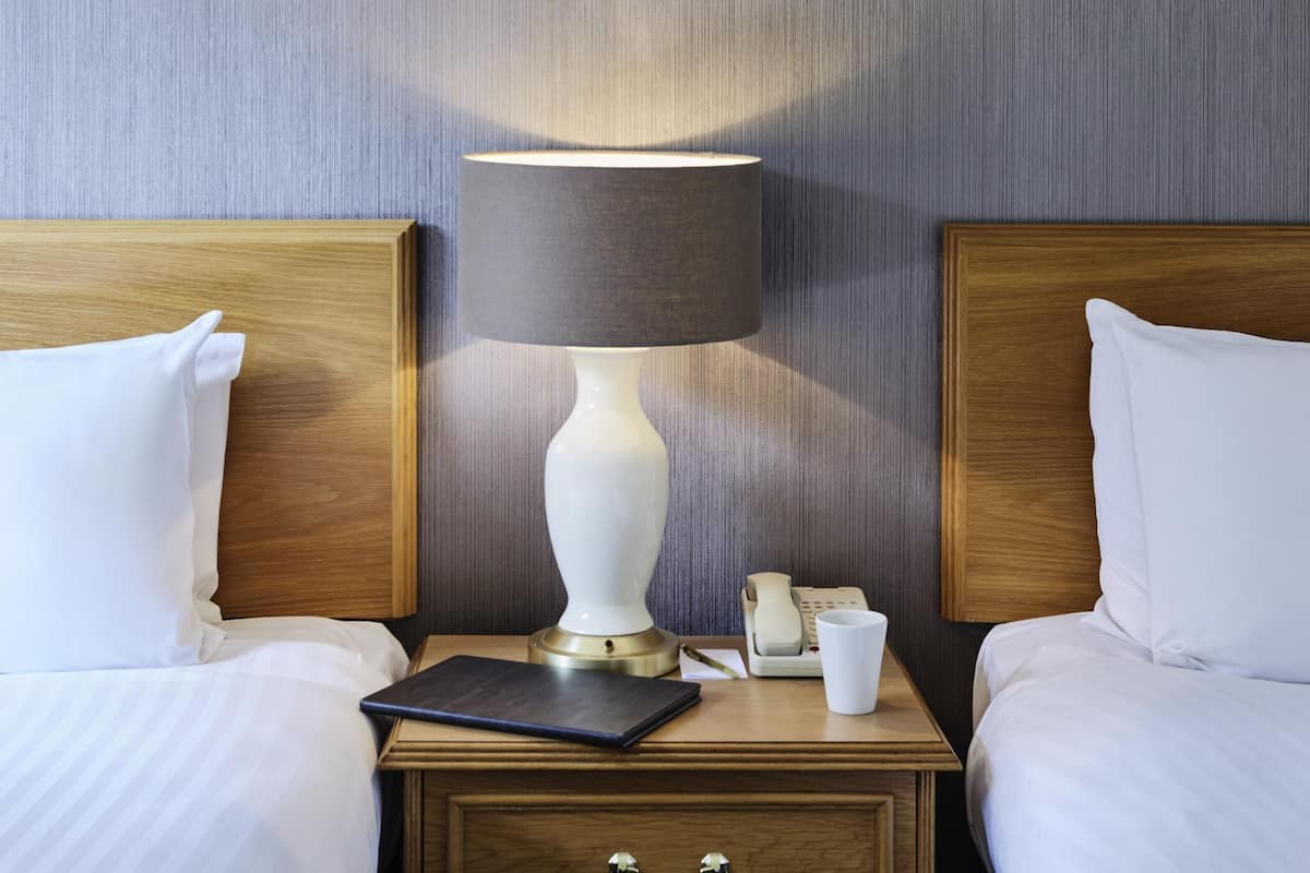 Quarto de hotel com cabeceira de madeira, luminária branca, mesa de cabeceira com telefone, xícara e caderno.