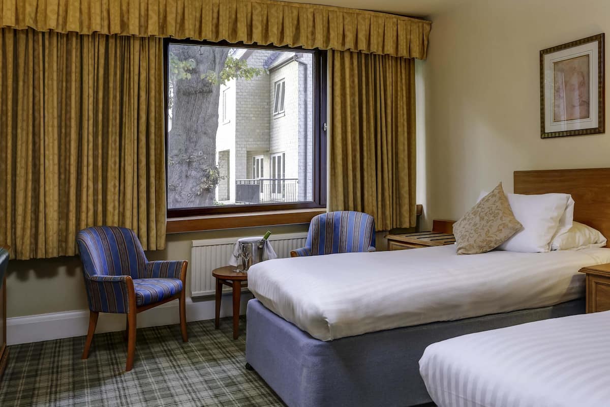 Ein Hotelzimmer mit Bett, zwei Stühlen, einem kleinen Tisch mit einem Glas und einem Fenster mit Vorhängen.