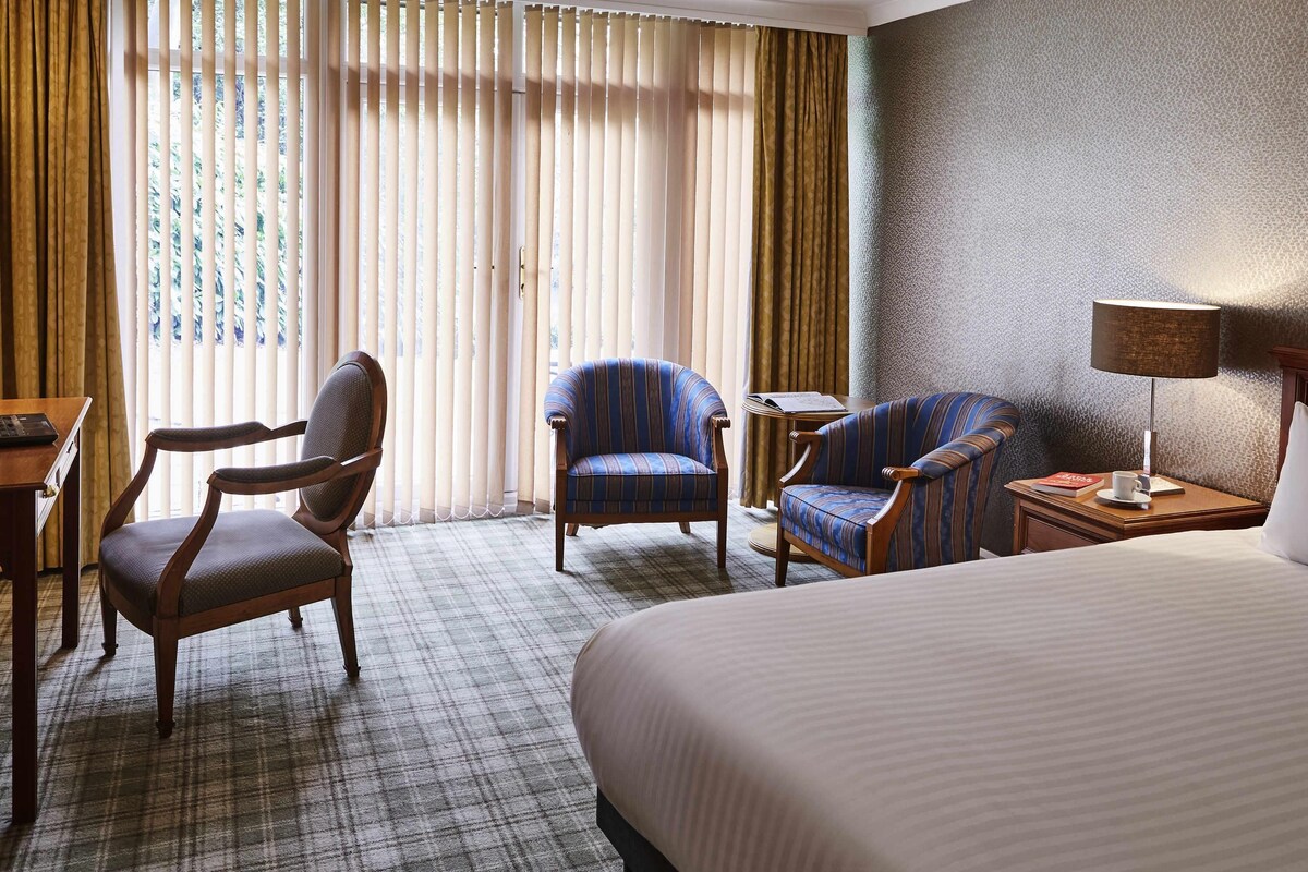 A hotel room with a large bed, two armchairs, a desk with a lamp, and a view of the outdoors through sheer curtains.