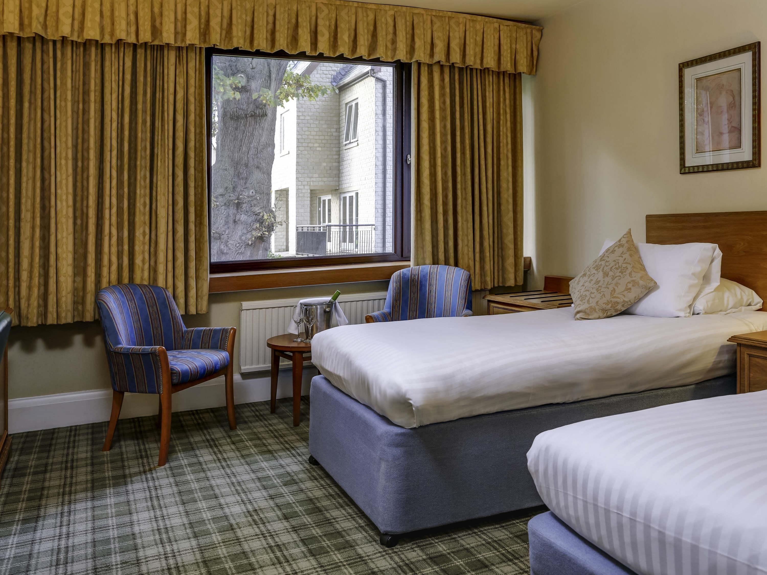 A hotel room with a bed, two chairs, a small table with a glass, and a window with curtains.