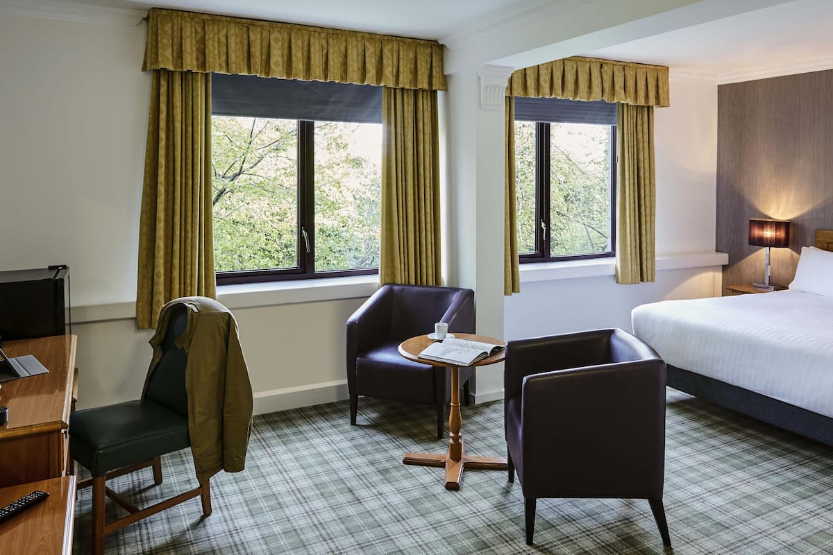 Ein Hotelzimmer mit einem Bett, zwei Stühlen, einem kleinen Tisch mit einem Buch, einem Schreibtisch mit einem Laptop und einem Fernseher.
