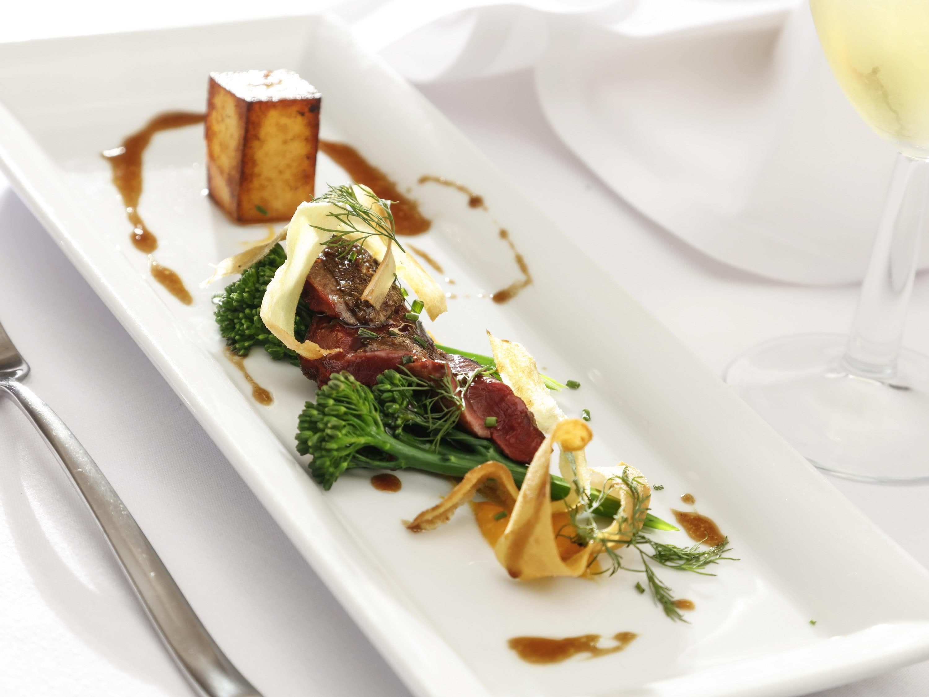 Un plato blanco rectangular con un guiso, una copa de vino blanco y un tenedor a la izquierda.