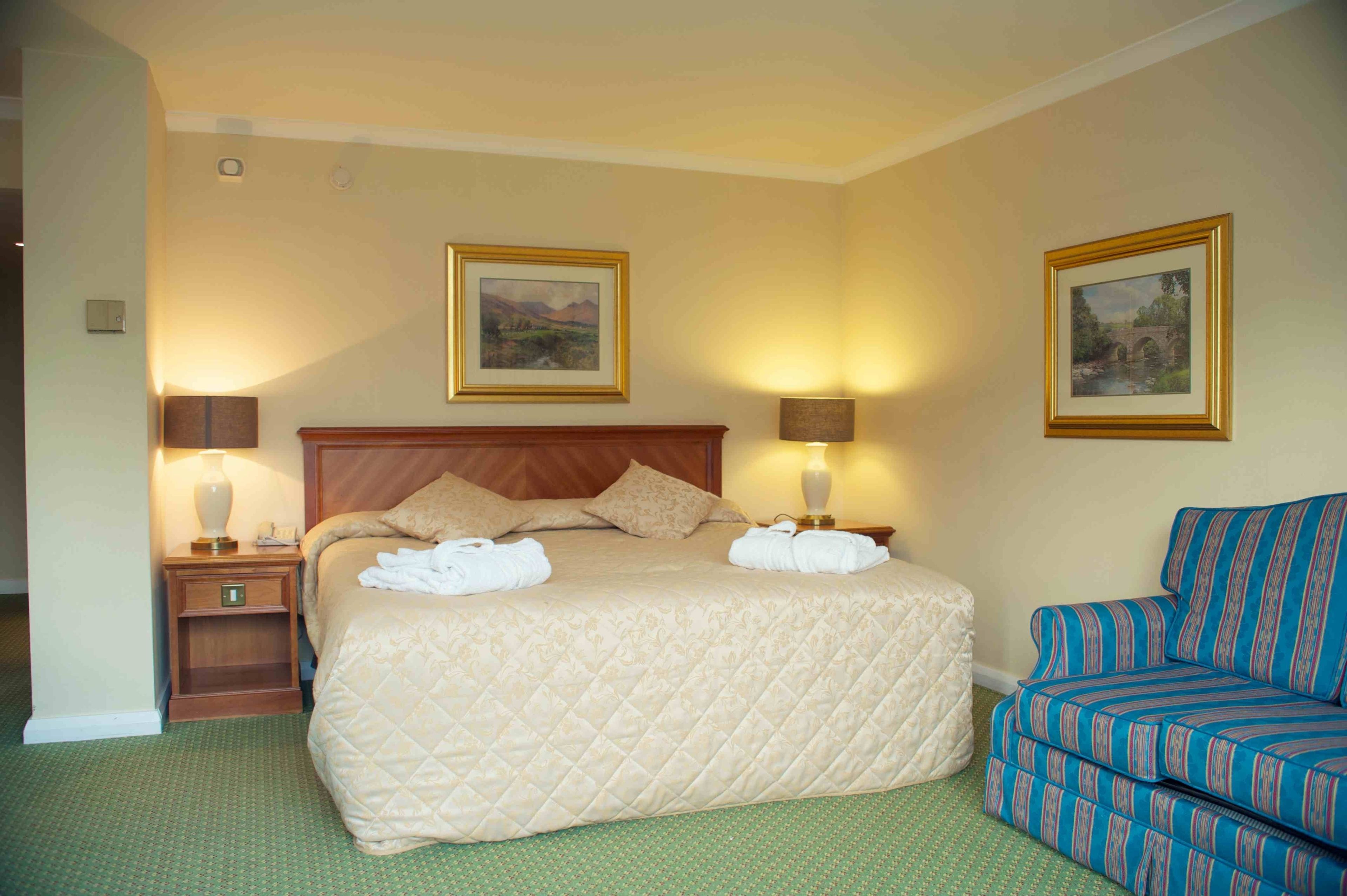 A hotel room with a large bed, two bedside lamps, a nightstand, a sofa, and two framed pictures on the walls.