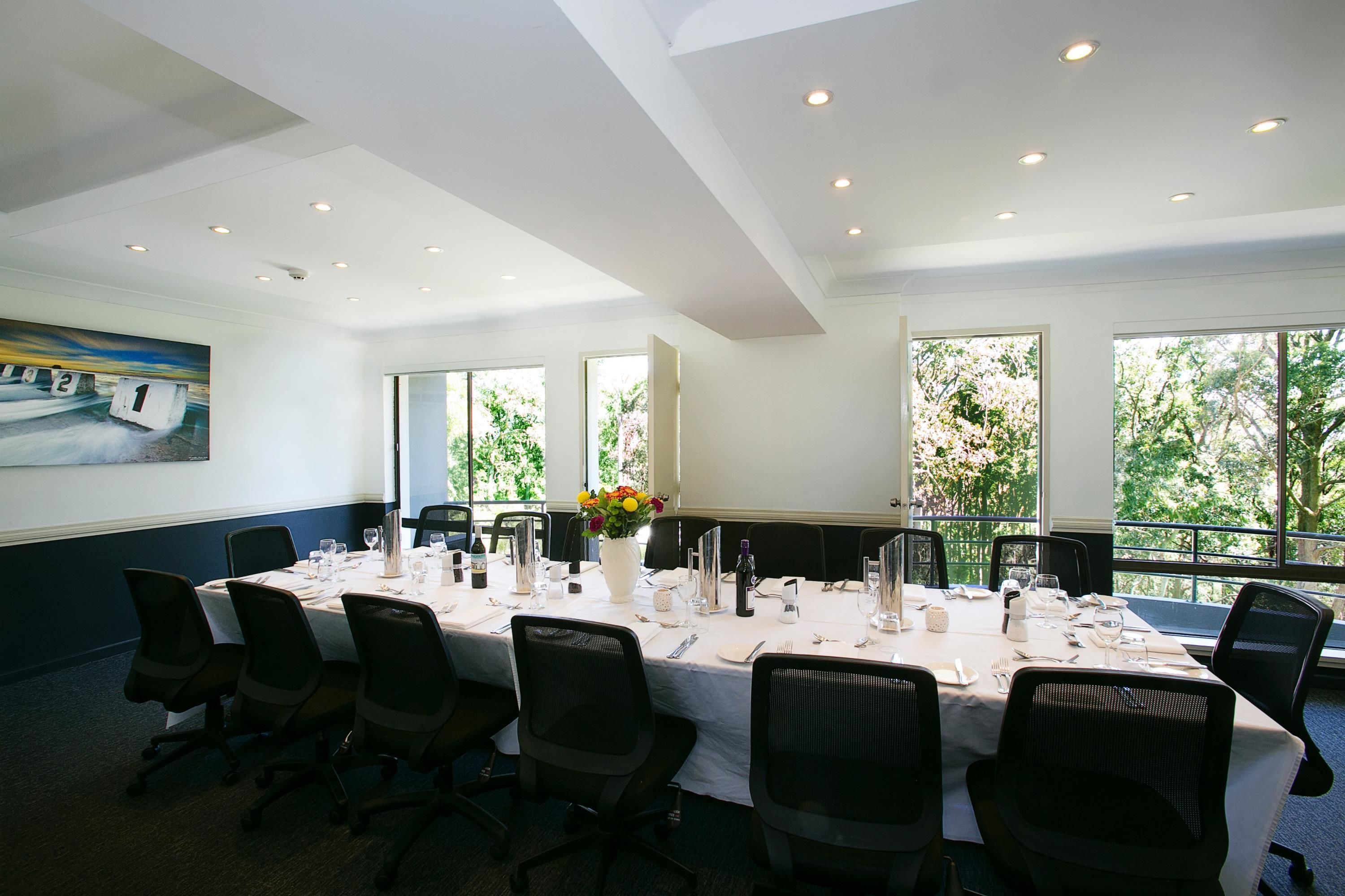 A conference room with a long table set for a meeting, large windows, and a painting on the wall.