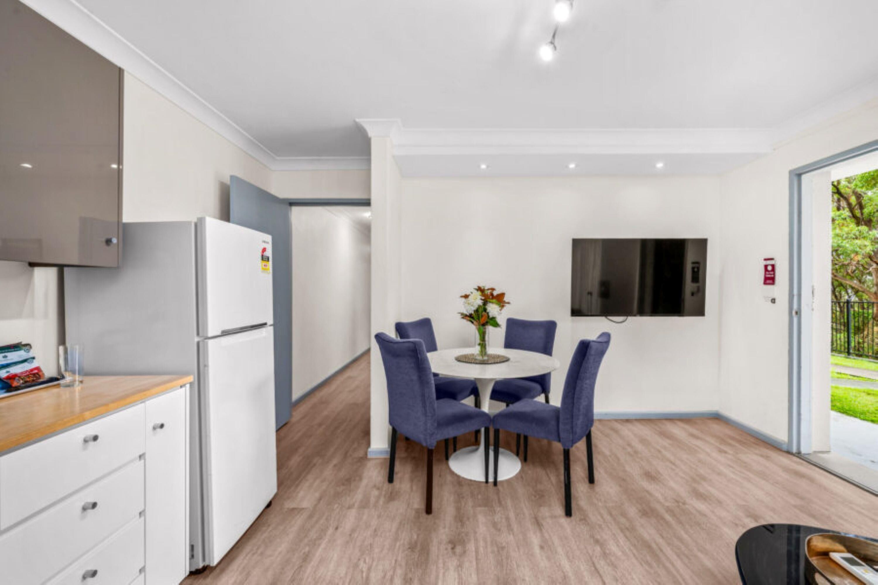 A modern kitchen with white cabinets, a built-in refrigerator, and a dining area with a round table and blue chairs.