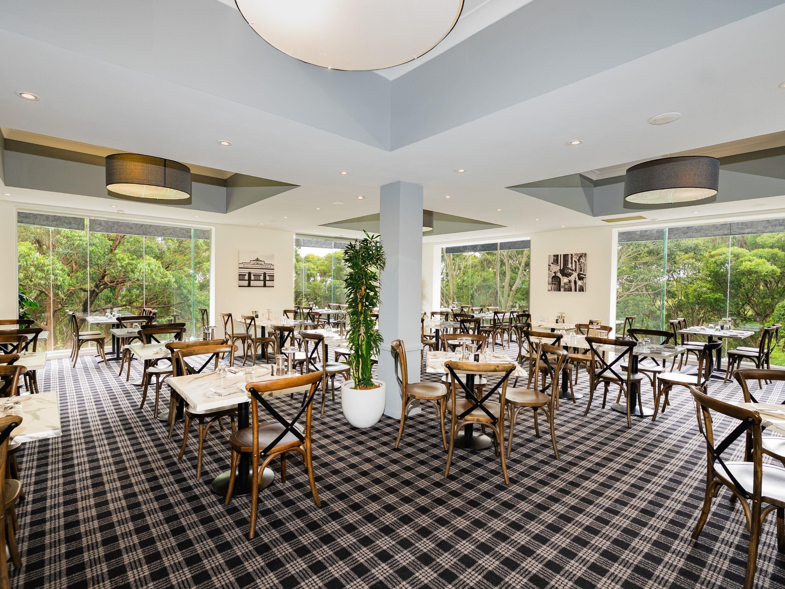 A spacious restaurant with a checkered floor, wooden chairs, and tables. Large windows offer a view of trees.
