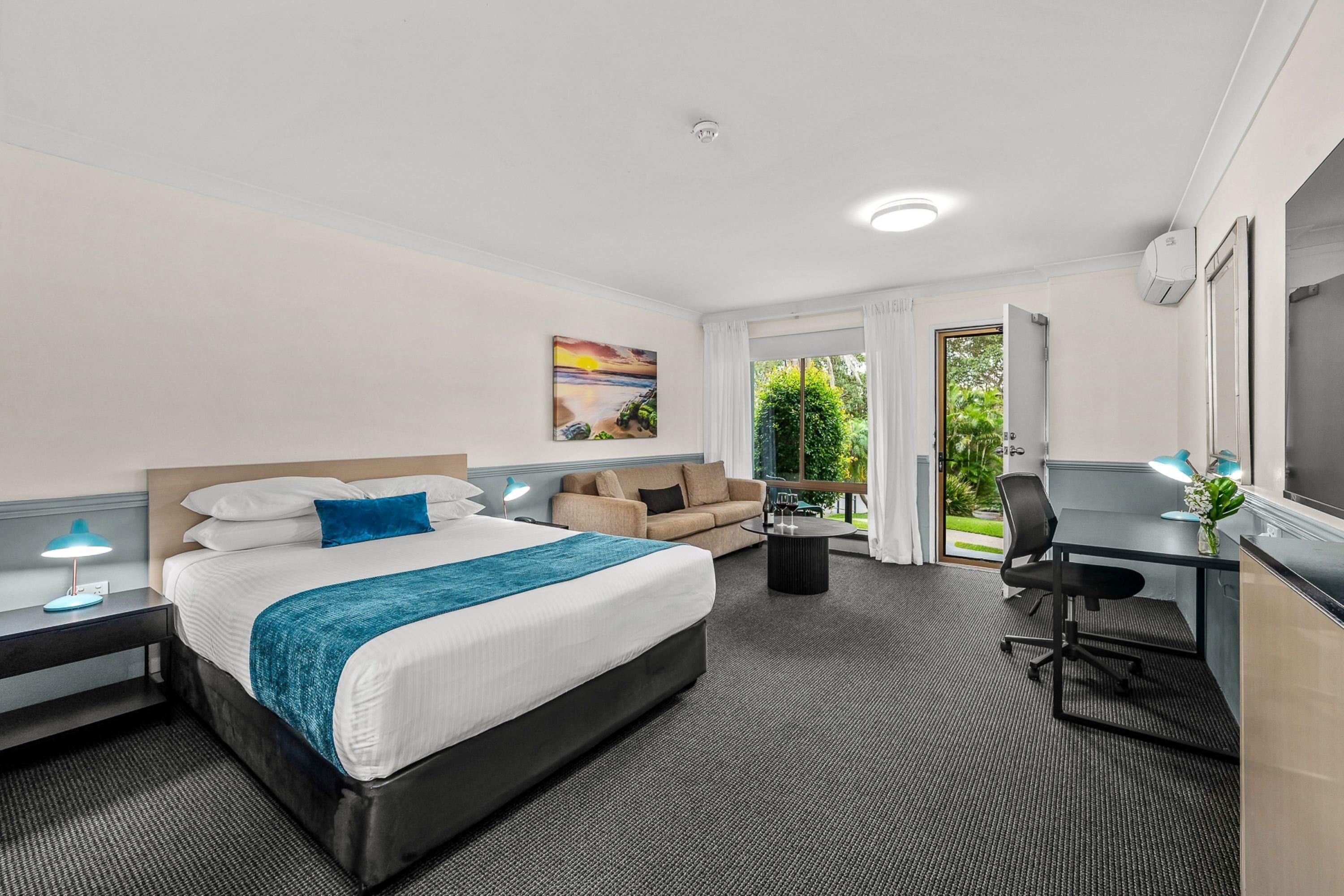 A hotel room with a bed, a desk, a chair, a sofa, a lamp, and a painting on the wall.