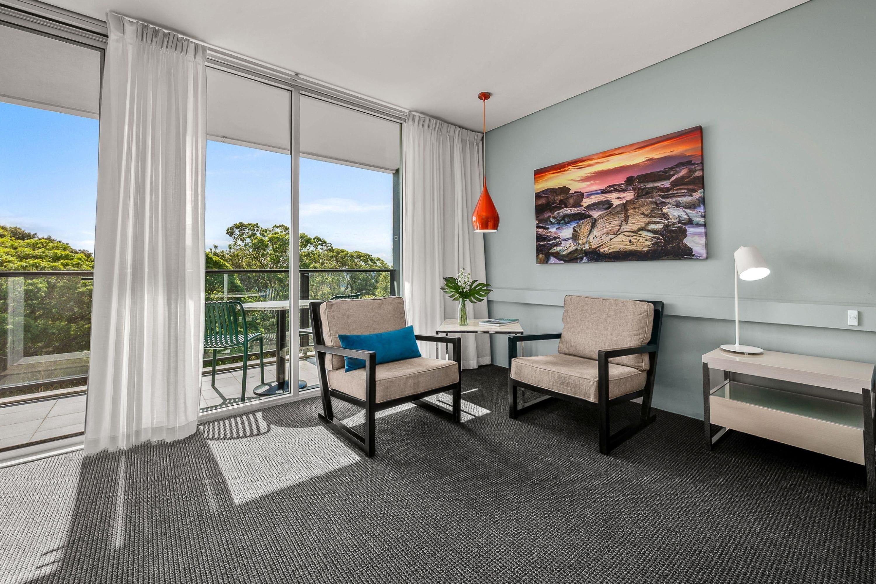 A modern room with a balcony, two chairs, a small table, a lamp, and a painting on the wall.