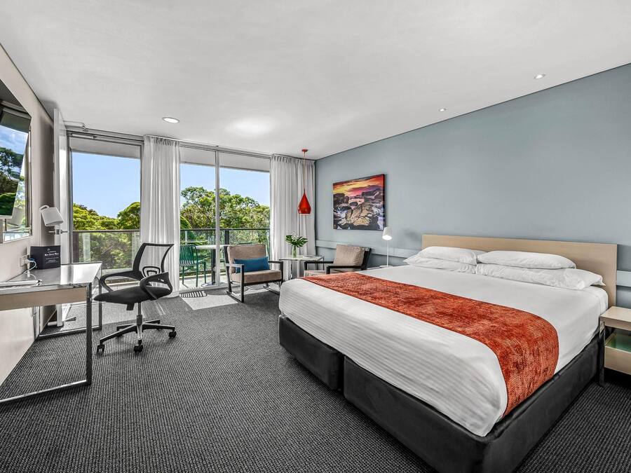 A modern hotel room with a large bed, a desk, a chair, a balcony with a view, and a painting on the wall.