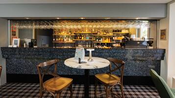 Uma área de bar com bancada de mármore, duas cadeiras de madeira, um suporte para menu e diversos frascos e copos em prateleiras.