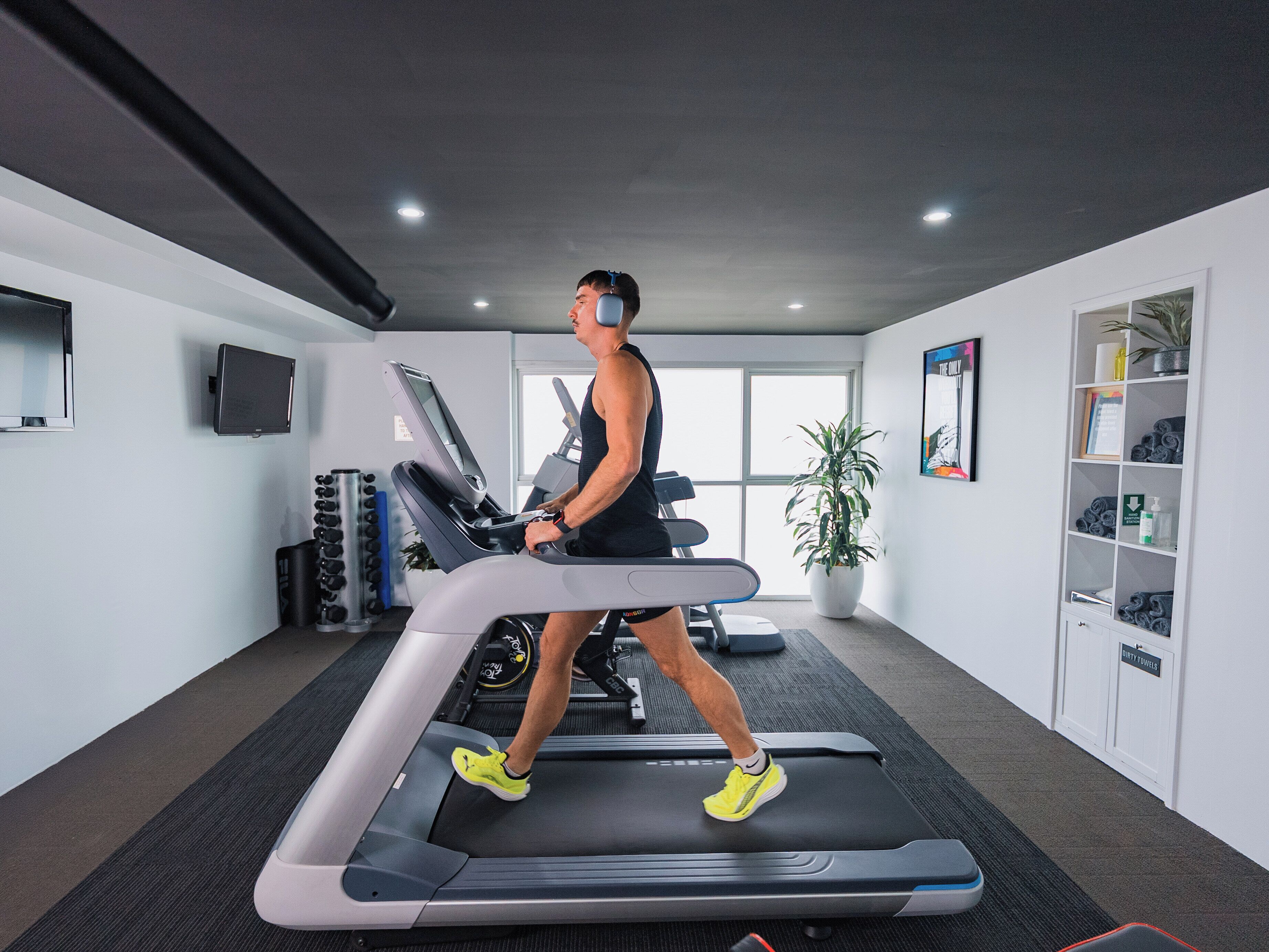 A man walking on a treadmill in a gym with a TV, exercise equipment, and a plant.
