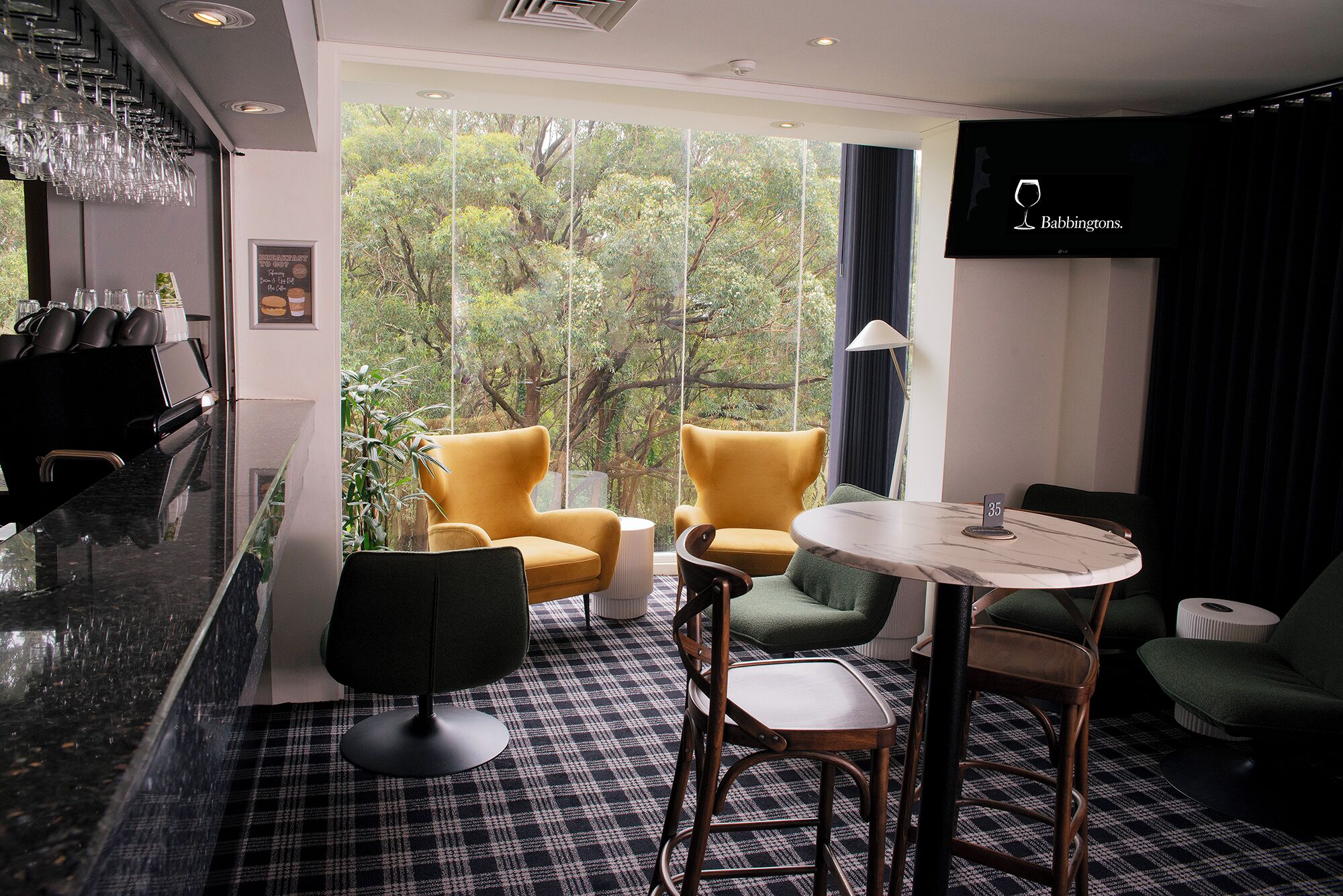 A modern room with a bar area, two yellow armchairs, a green armchair, a round table, and a view of trees through large windows.