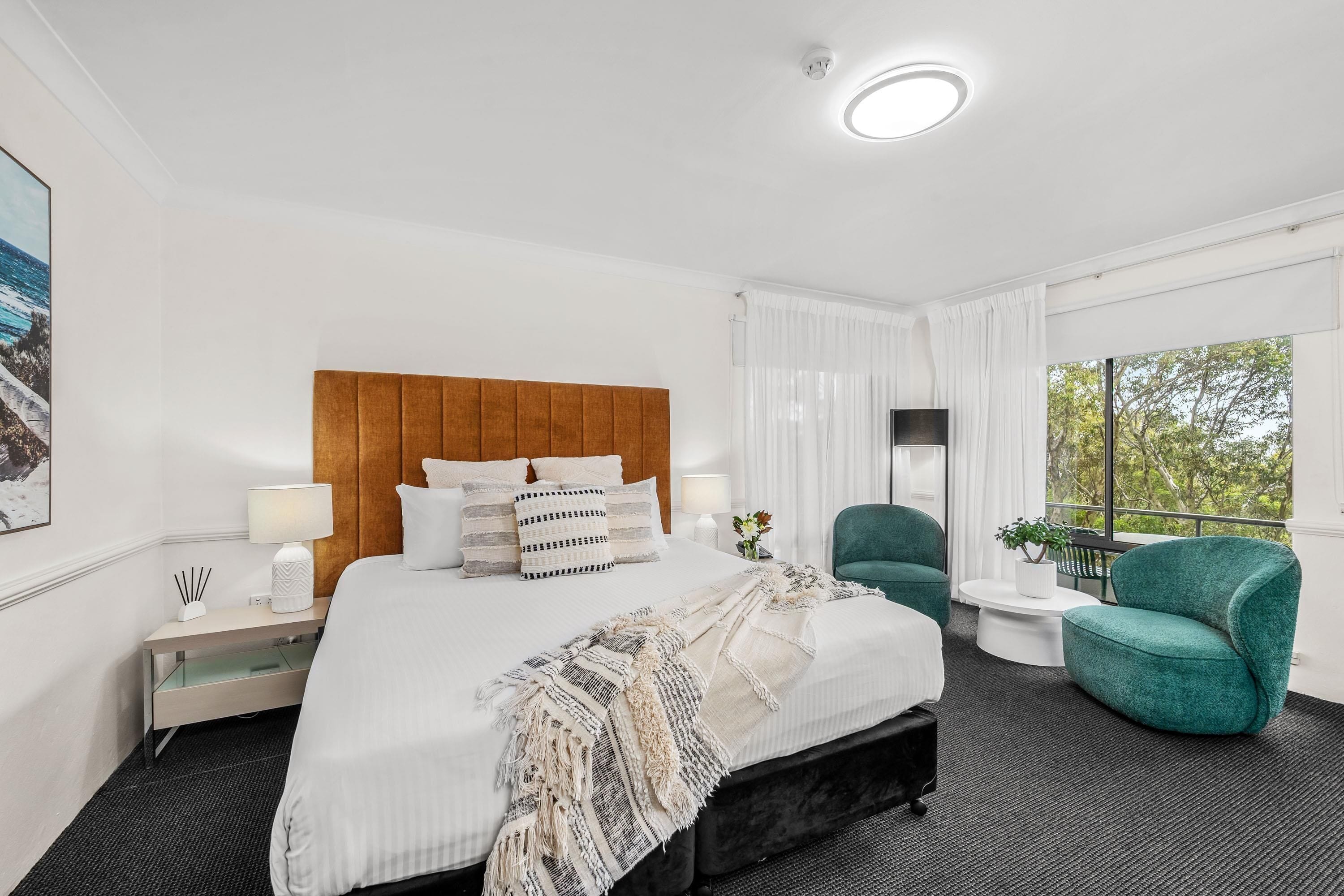 A hotel room with a large bed, a wooden headboard, a green armchair, a small side table, and a window with a view of trees.