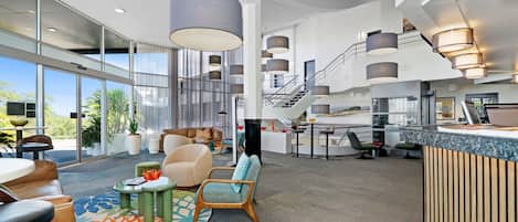 Un amplio vestíbulo de hotel con techo alto, ventanas grandes y una escalera central.