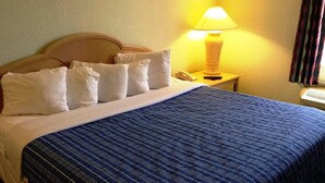 A neatly made bed with a blue striped bedspread and white pillows, a bedside lamp, and a telephone on a wooden nightstand. - Cocoa Beach Suites (Cocoa Beach)