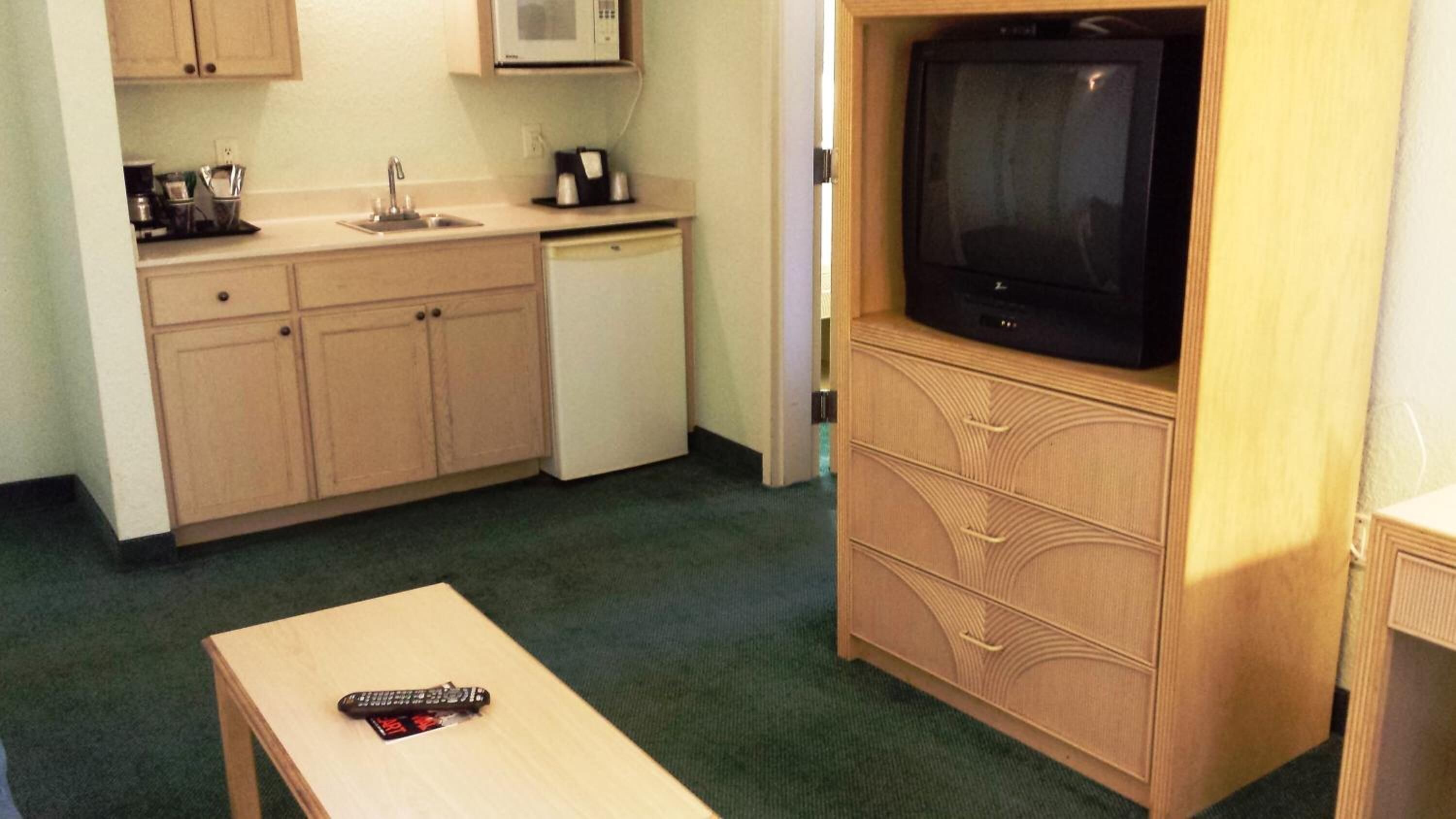 A compact kitchenette with a microwave, sink, and coffee maker. A television is mounted on a wooden cabinet, and a remote control is on a small table.