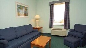 A room with a blue sofa, a wooden coffee table, and a smaller blue armchair. There is a window with curtains, a lamp on a side table, and a painting on the wall. - Cocoa Beach Suites (Cocoa Beach)