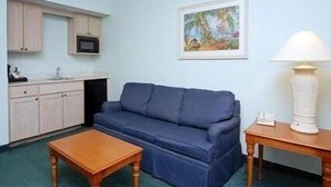 A compact living space with a blue sofa, wooden coffee table, kitchenette with cabinets, and a painting on the wall. - Cocoa Beach Suites (Cocoa Beach)