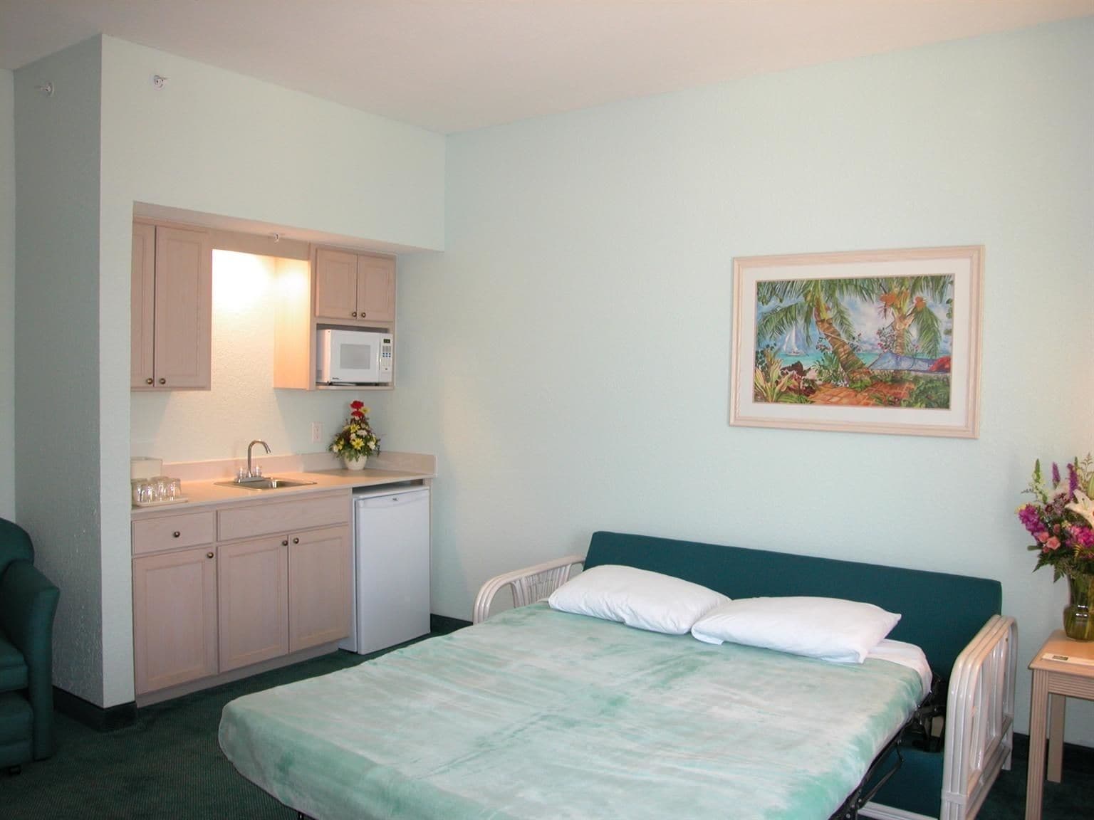 Een slaapkamer met een bed, een klein keukentje, een stoel en een schilderij aan de muur.