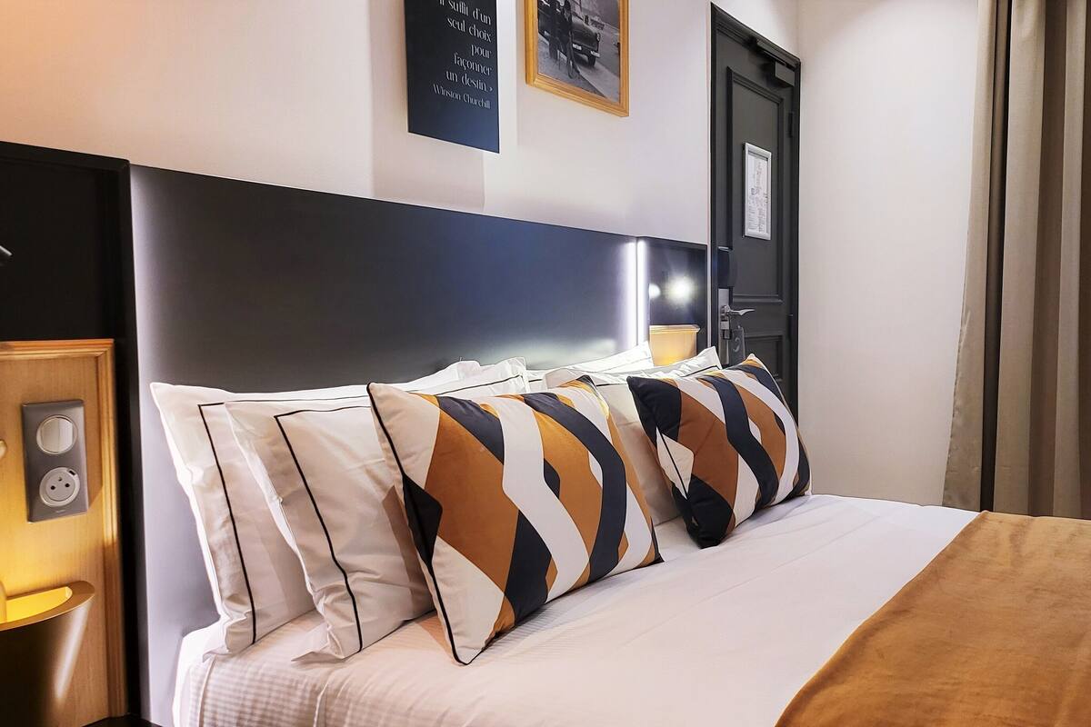 Habitación de hotel con cama, mesita de noche, un reloj en la pared y una puerta con manija.