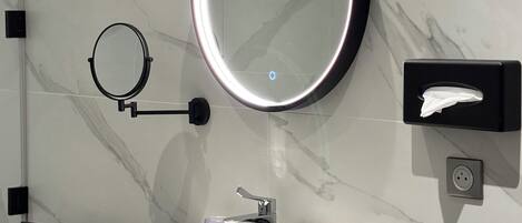 A modern bathroom with a marble wall, a round mirror with a light, a sink, a towel rack, and a hand dryer.