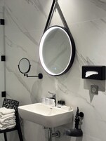A modern bathroom with a marble wall, a round mirror with a light, a sink, a towel rack, and a hand dryer.
