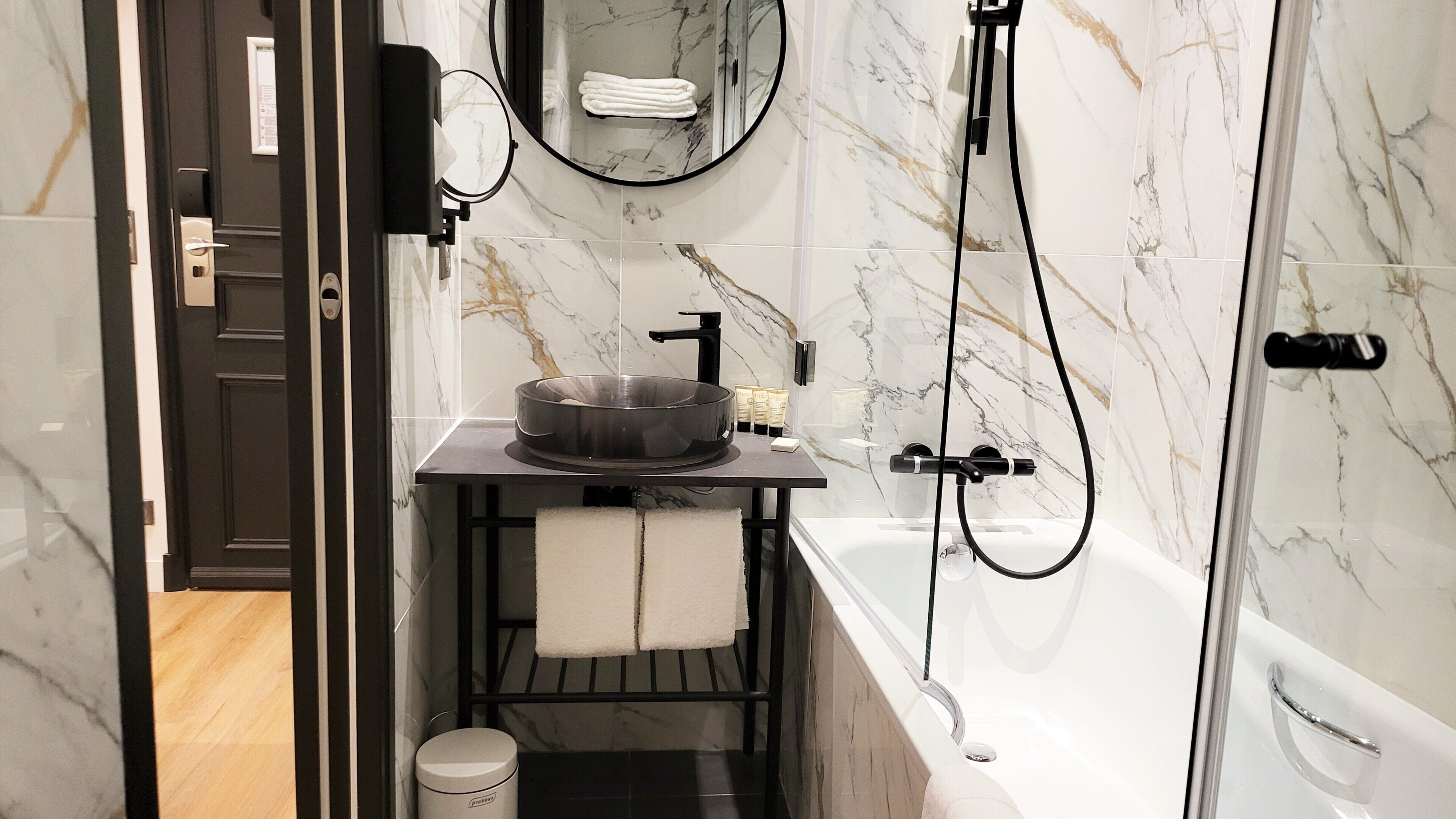 Un baño moderno con salpicadero de mármol, un lavabo negro, un espejo grande, un cabezal de ducha manual y una bañera.