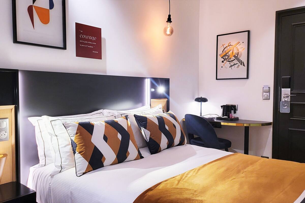 Una habitación de hotel moderna con una cama grande, un escritorio y una silla. Hay una cita enmarcada en la pared y una lámpara sobre el escritorio.